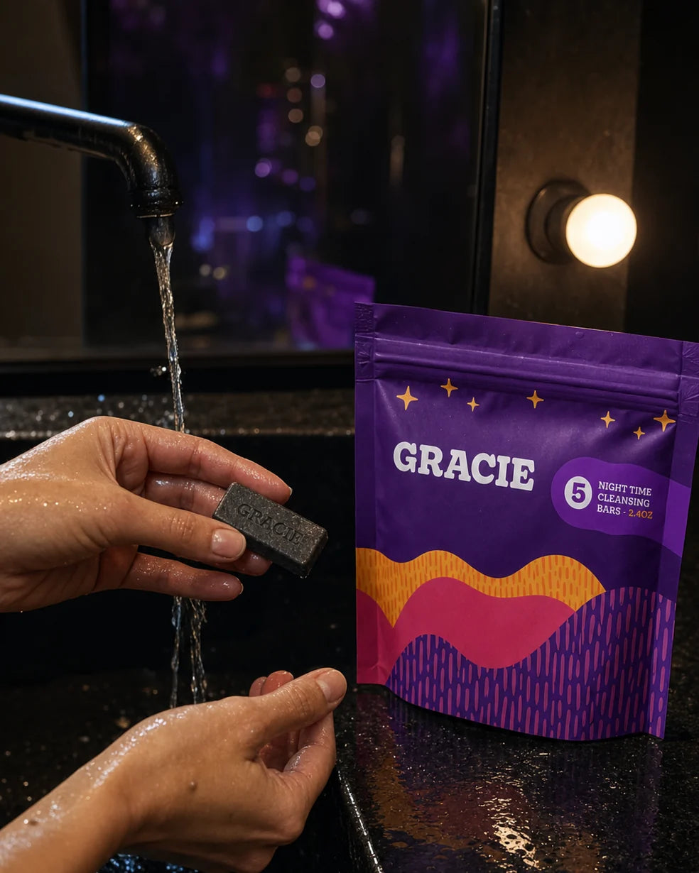 A hand holding the charcoal Gracie Evening mini bar beside its violet pouch at a backstage sink