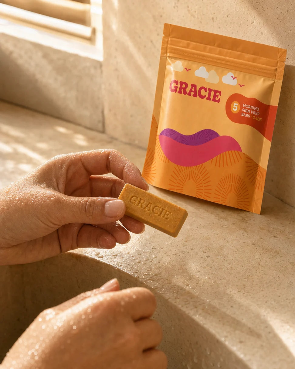 A hand holding the gold Gracie Morning mini bar beside its pouch at a sunlit sink